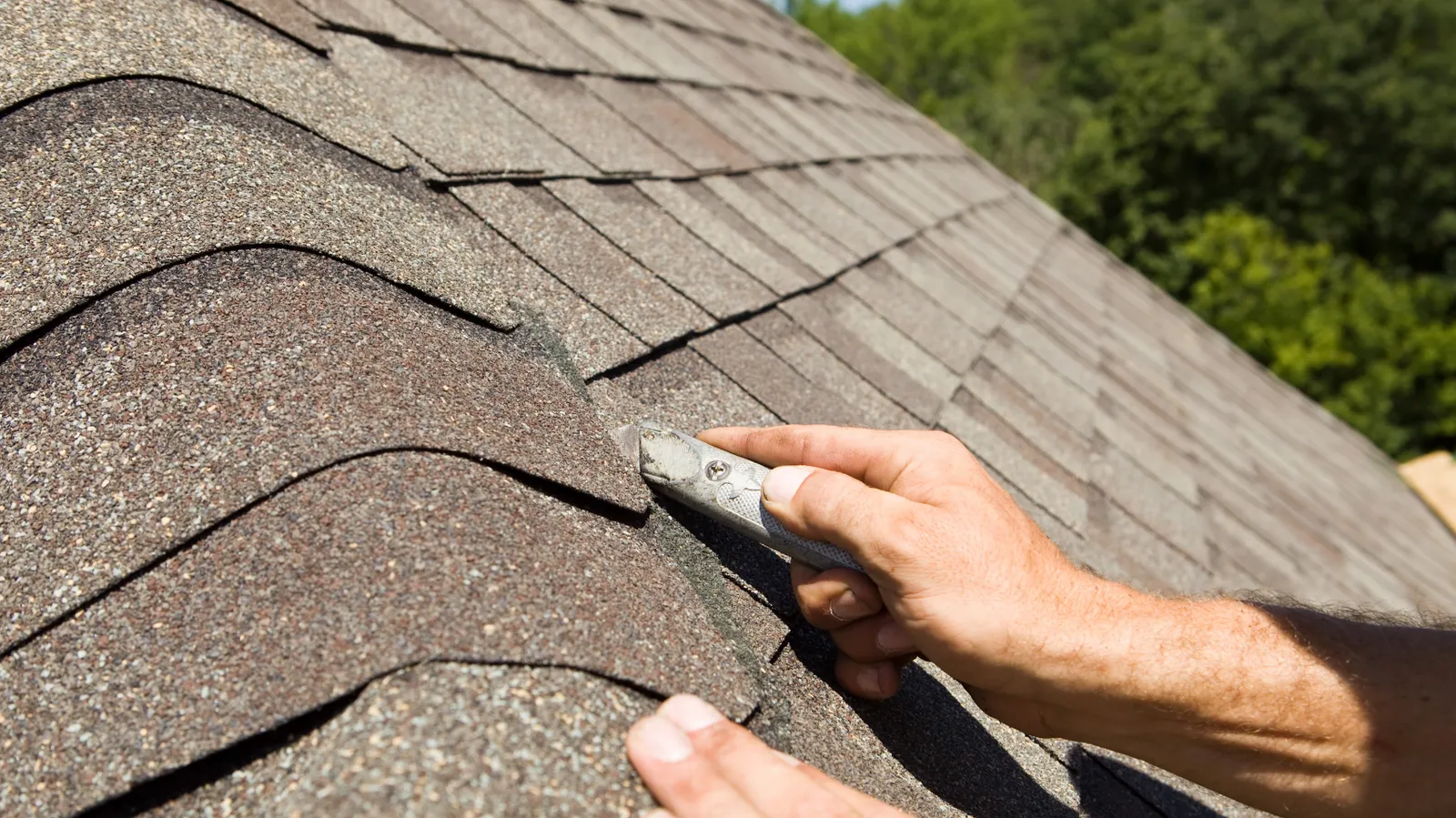 A roofer trimming a ridge cap shingle with a utility knife